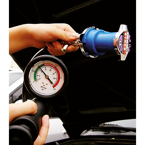 Person hält ein Kühlsystem-Prüfgerät über dem Motorraum eines Autos, ein Manometer zeigt den Druck an.